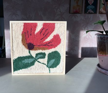 Zoë Handgetuft wolschilderij met crème kleurige achtergrond. Er staat een grote roze bloem op met een groene steel en groene bladeren.